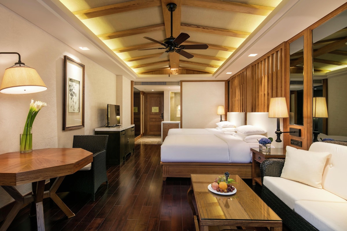 A hotel room with a bed, a sofa, a dining table, a TV, and a ceiling fan.