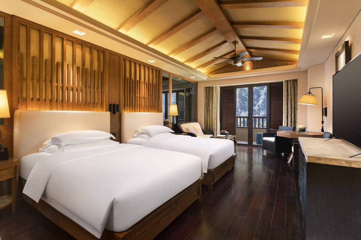 Una habitación de hotel con dos camas, un ventilador de techo, un ventanal grande y techo de madera.