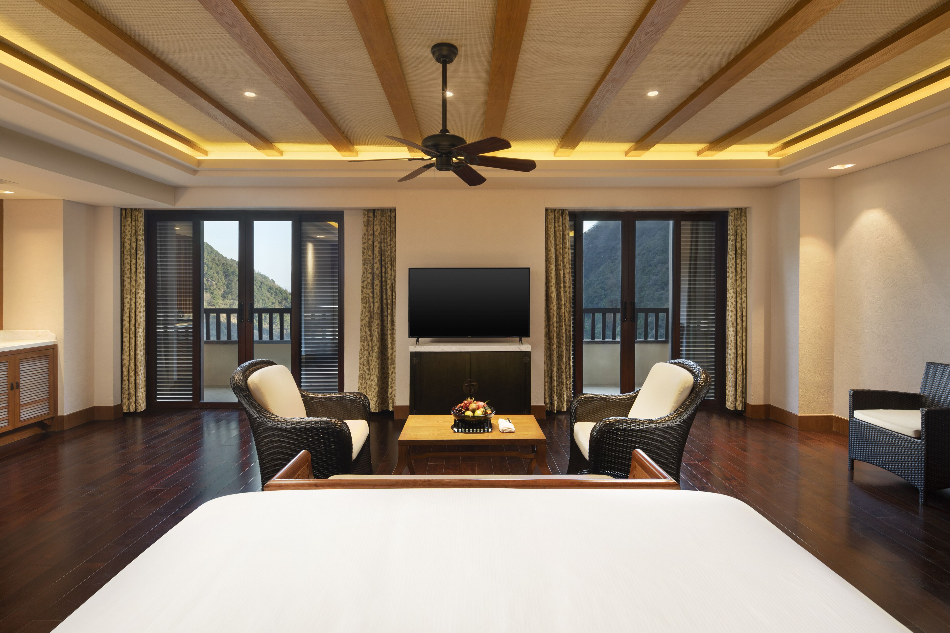 Amplia habitación de hotel con una cama grande, televisión de pantalla plana, ventilador de techo y vistas a la montaña a través de las ventanas.