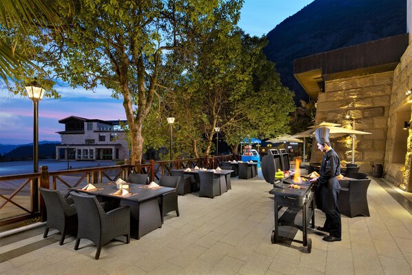 An outdoor dining area with tables and chairs, a chef preparing food, and a view of a mountain and hotel in the background. - Hilton Sanqingshan Resort (Shangrao)