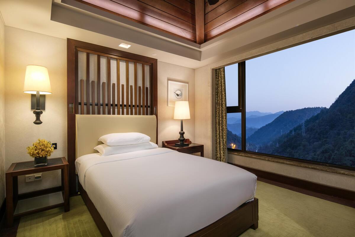 Una habitación de hotel con una cama, dos mesitas de noche, una lámpara, un reloj de pared y un amplio ventanal con vistas a la montaña.