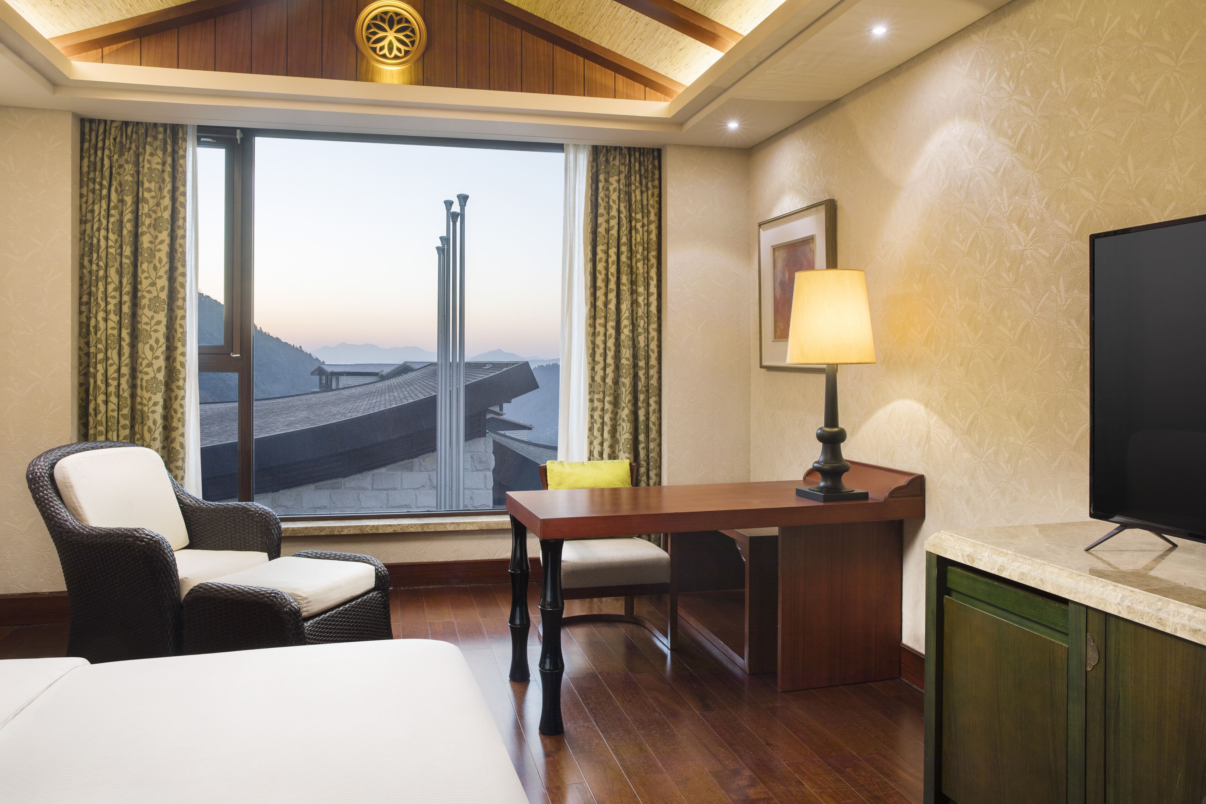 Habitación de hotel con cama, escritorio, silla, televisor y ventana con vista a un puente y montañas.