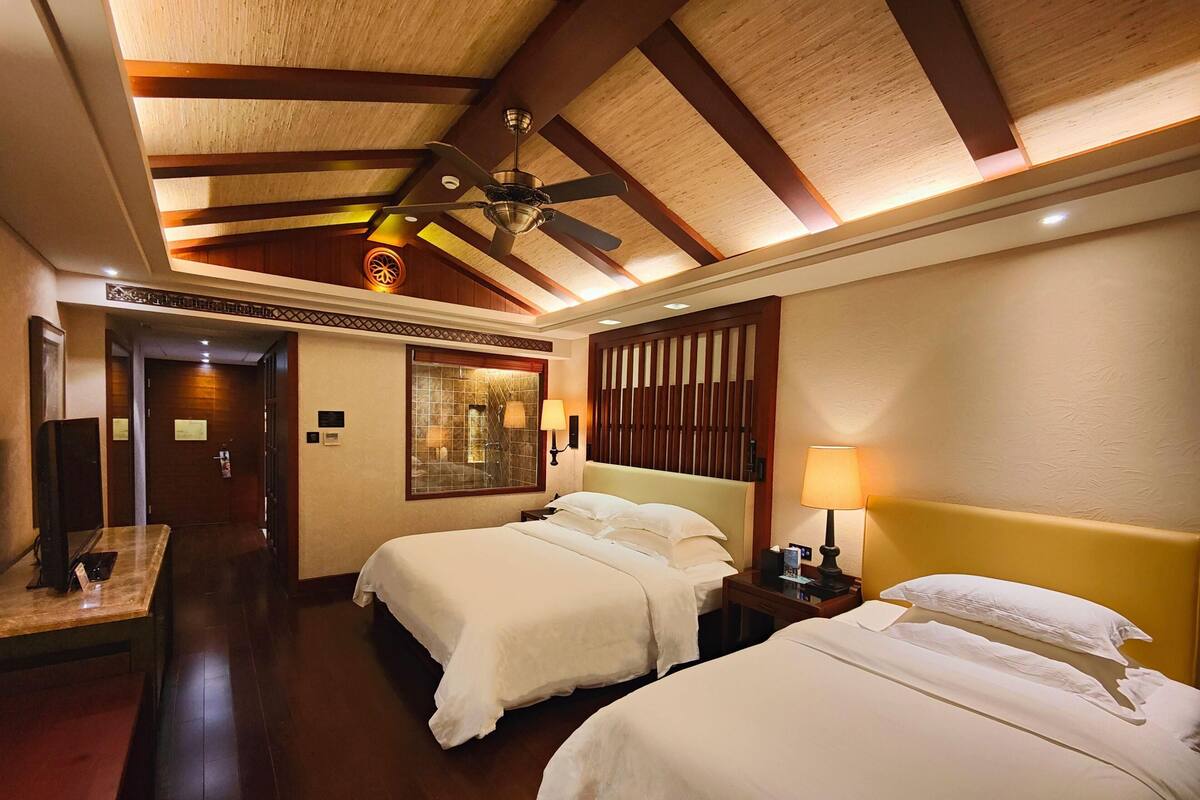 A hotel room with two beds, a ceiling fan, a large mirror, a desk, and a door leading to a hallway.