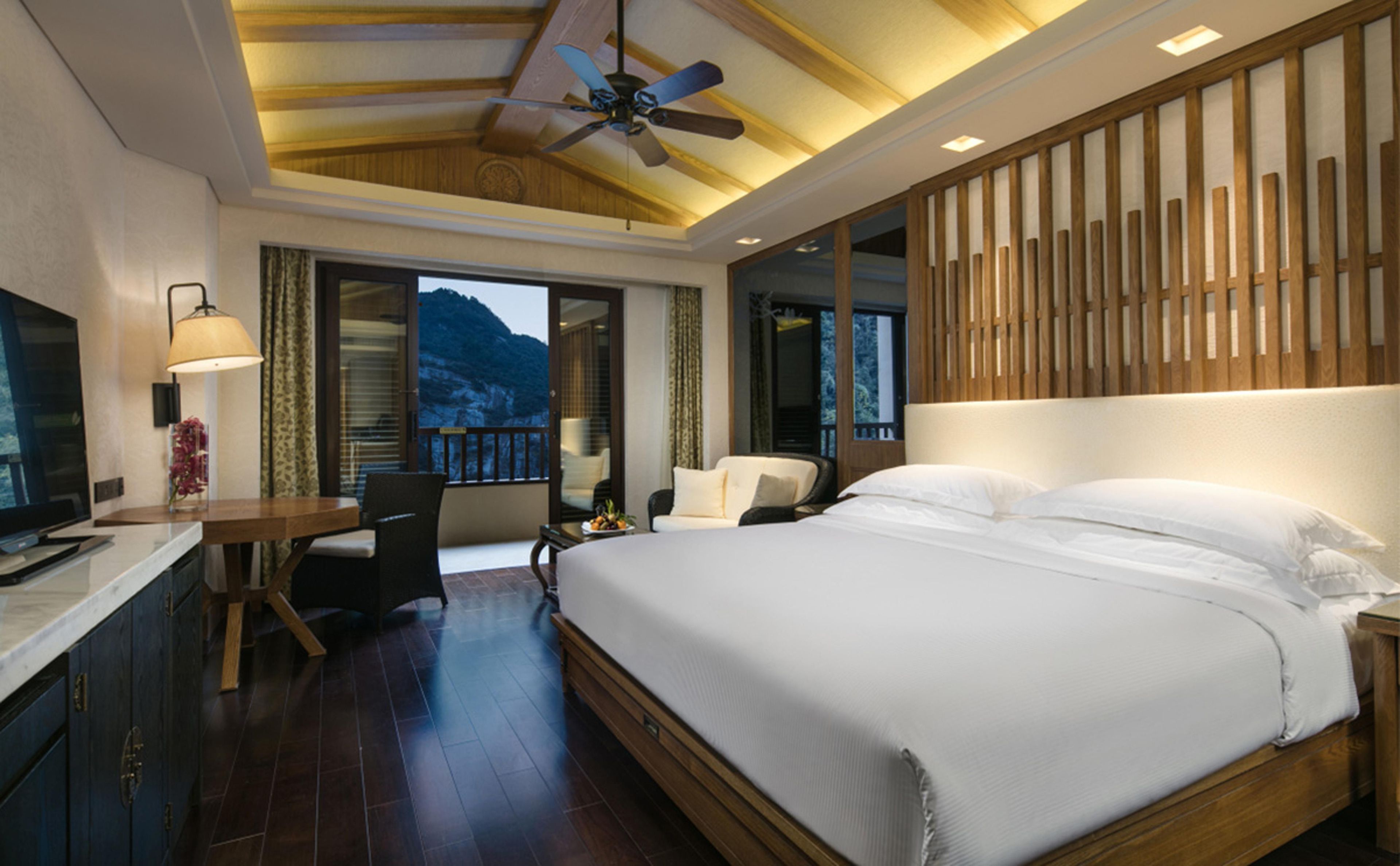 Habitación de hotel con una cama grande, un escritorio con una silla, un televisor y un balcón con vistas a las montañas.