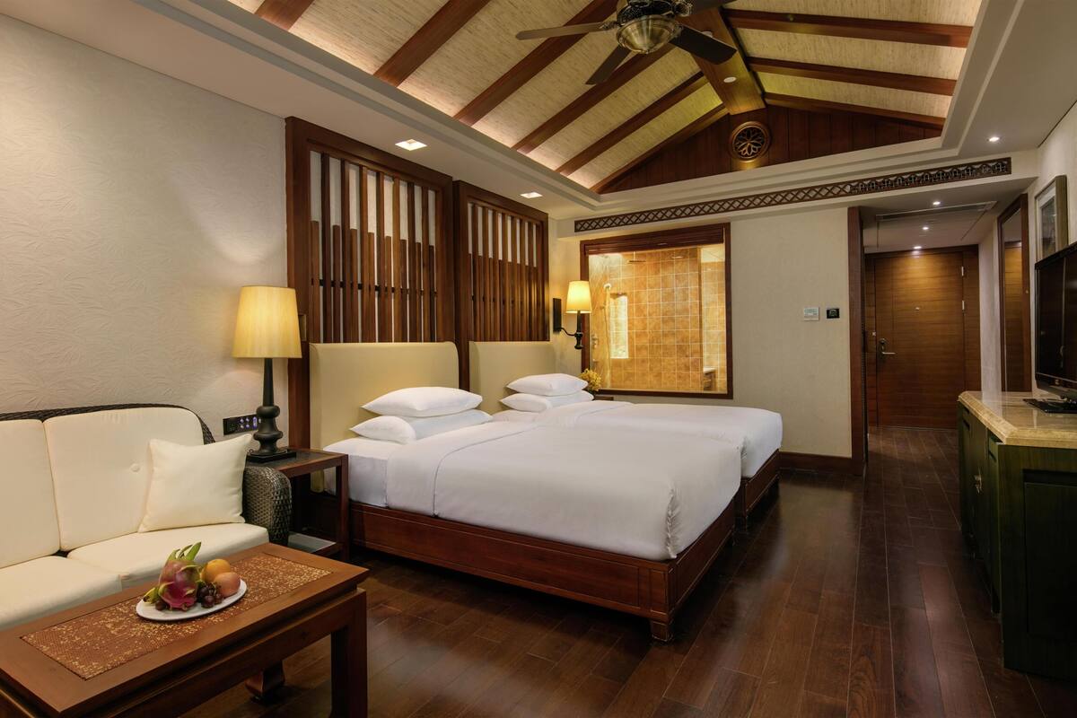Habitación de hotel con techo de madera, cama con ropa blanca, sofá, una mesita con fruta y televisor.