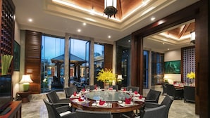 Uma ampla área de jantar com uma mesa redonda posta para refeição, rodeada por cadeiras de vime e grandes janelas com vista para o exterior.