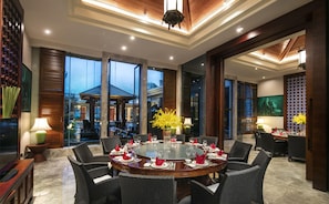 Un comedor bien iluminado con una mesa redonda preparada para una comida, rodeada de sillas de mimbre y grandes ventanas que ofrecen vistas al exterior.