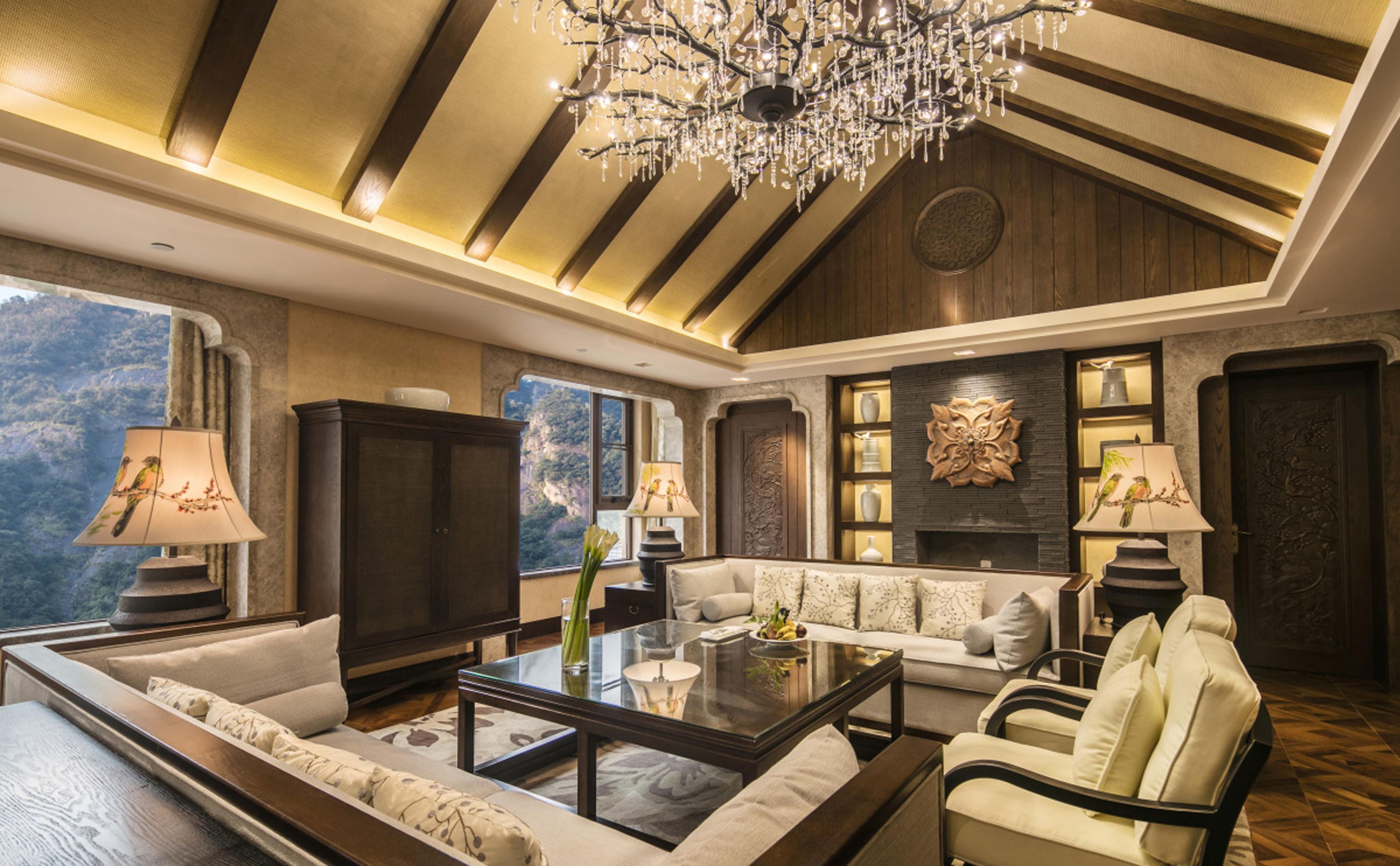 A spacious living room with a high ceiling, a chandelier, and a fireplace. There are multiple seating areas and a large window offering a view of the outdoors.