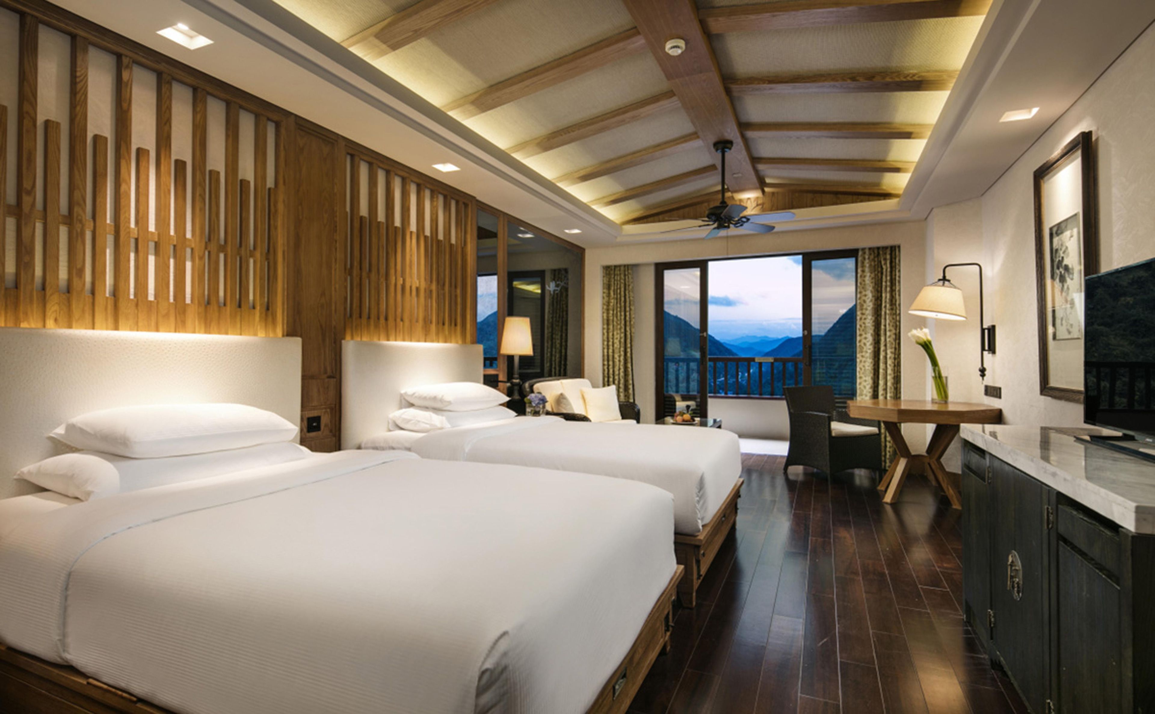A hotel room with two beds, a desk, a chair, a TV, and a view of mountains.