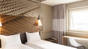 A hotel room with a bed, a desk, a chair, a telephone, and a window with curtains. - ibis De Haan (De Haan)