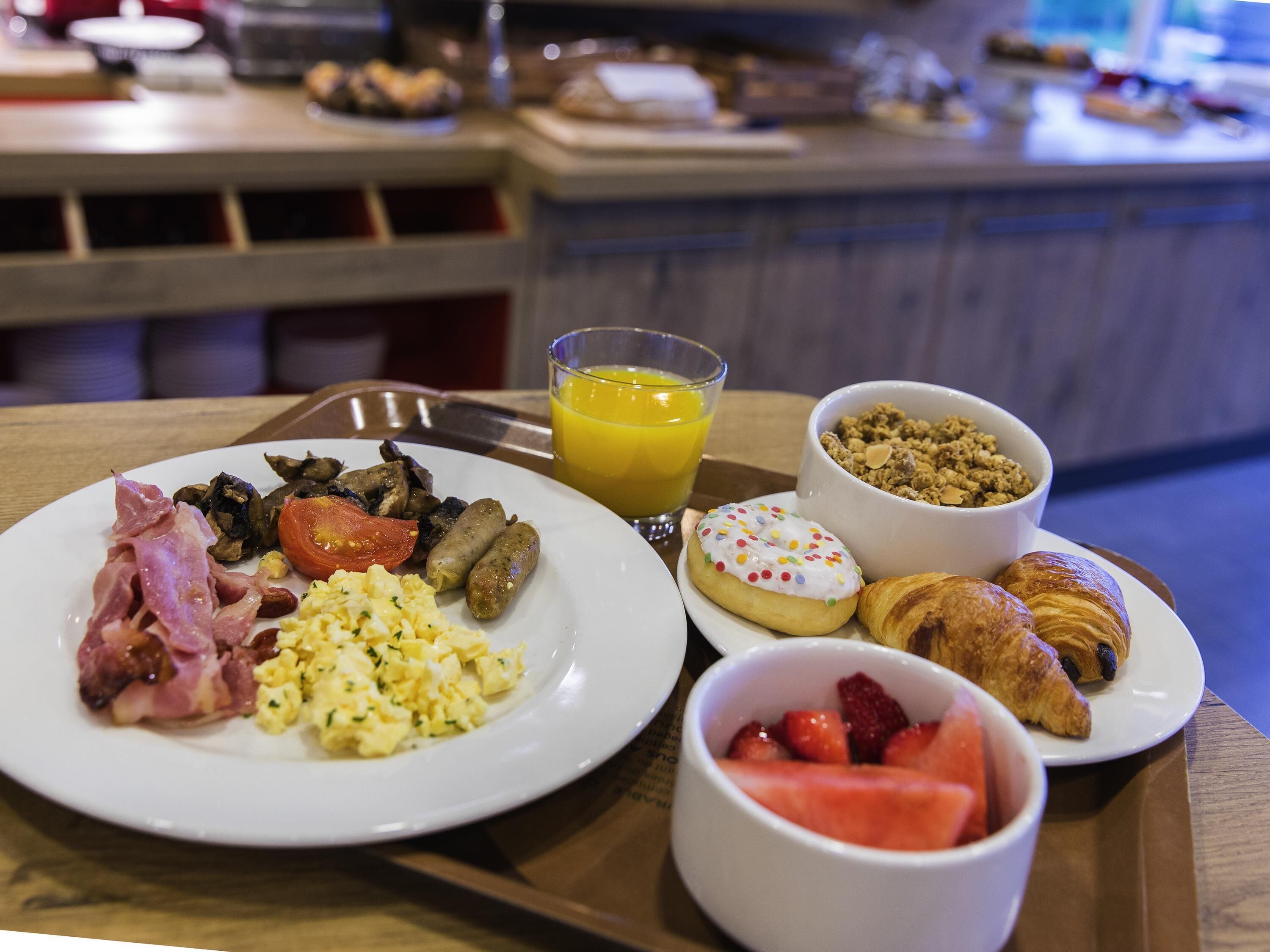 Un plateau de petit-déjeuner comprenant des œufs brouillés, des saucisses, du bacon, des champignons, une tomate, un beignet aux vermicelles, un croissant et un bol de fraises.