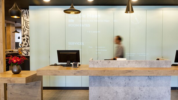 A hotel reception desk with a digital screen displaying room rates and services. - ibis De Haan (De Haan)