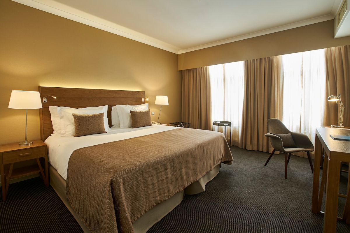 A hotel room with a large bed, two bedside tables with lamps, a desk with a chair, and a large window with curtains.