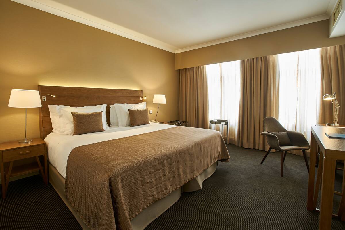 A hotel room with a large bed, two bedside tables with lamps, a desk with a chair, and a large window with curtains.