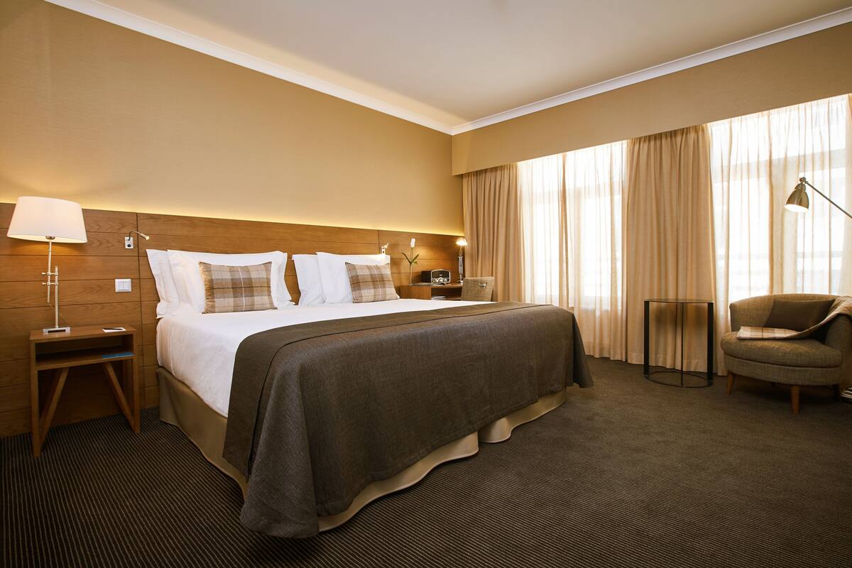 A hotel room with a large bed, a bedside table with a lamp, a chair, and a view of the outside through curtains.