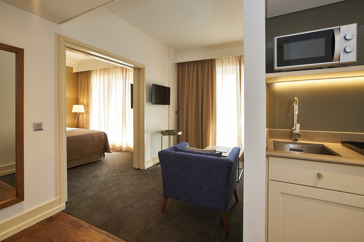 A hotel room with a bed, a sitting area with a chair, a TV mounted on the wall, a microwave, and a sink.