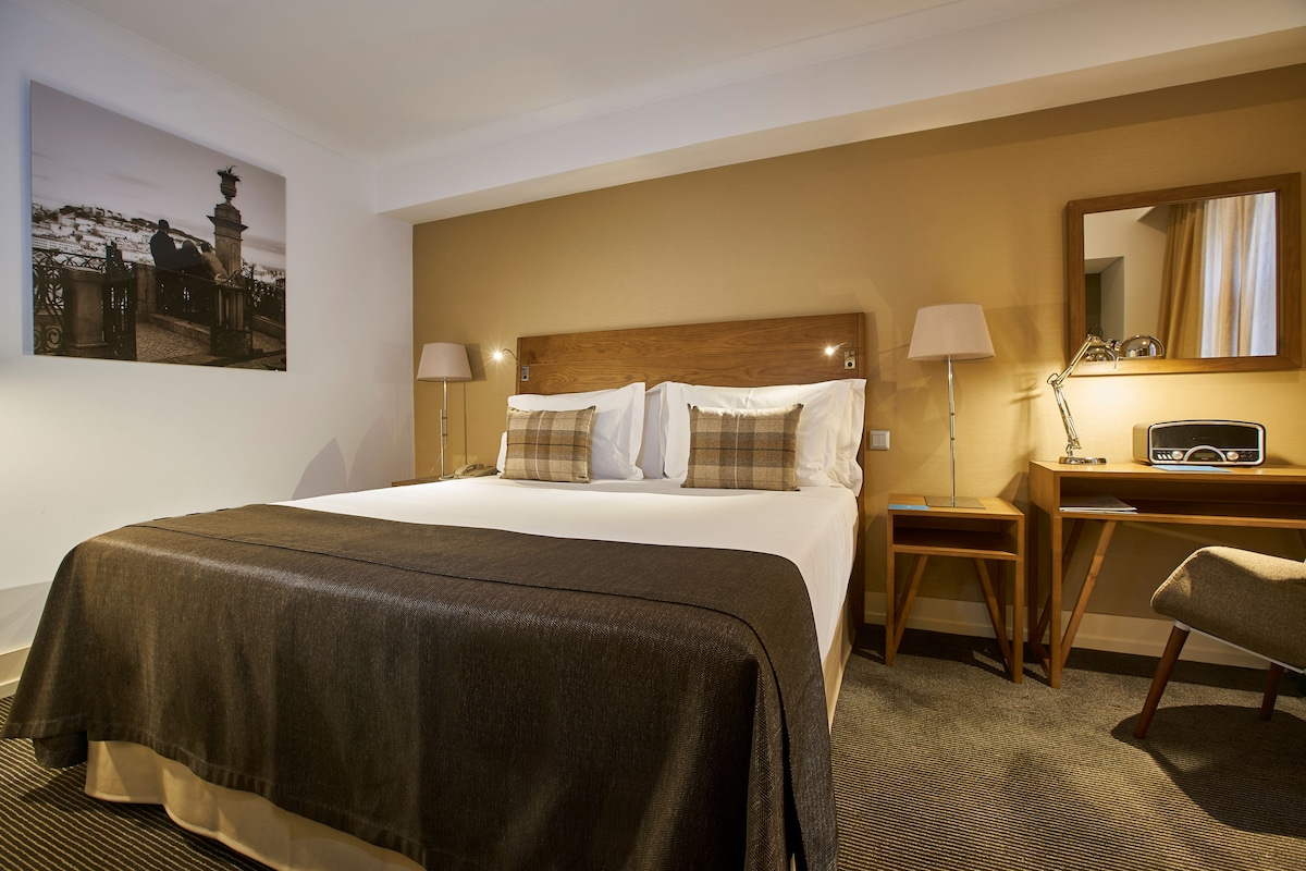 A hotel room with a large bed, two bedside tables, a desk, a chair, a lamp, and a painting on the wall.