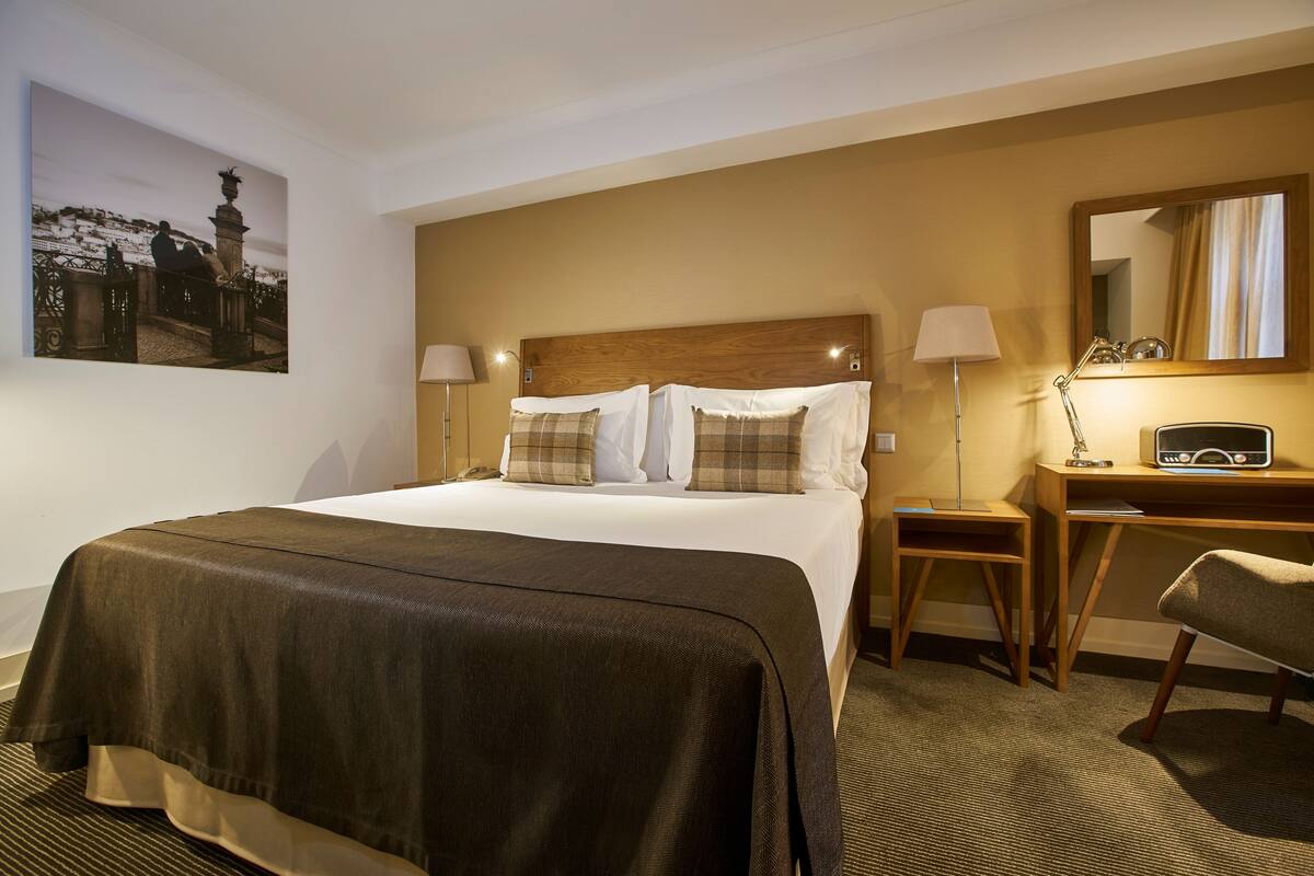 A hotel room with a large bed, two bedside tables, a desk, a chair, a lamp, and a painting on the wall.