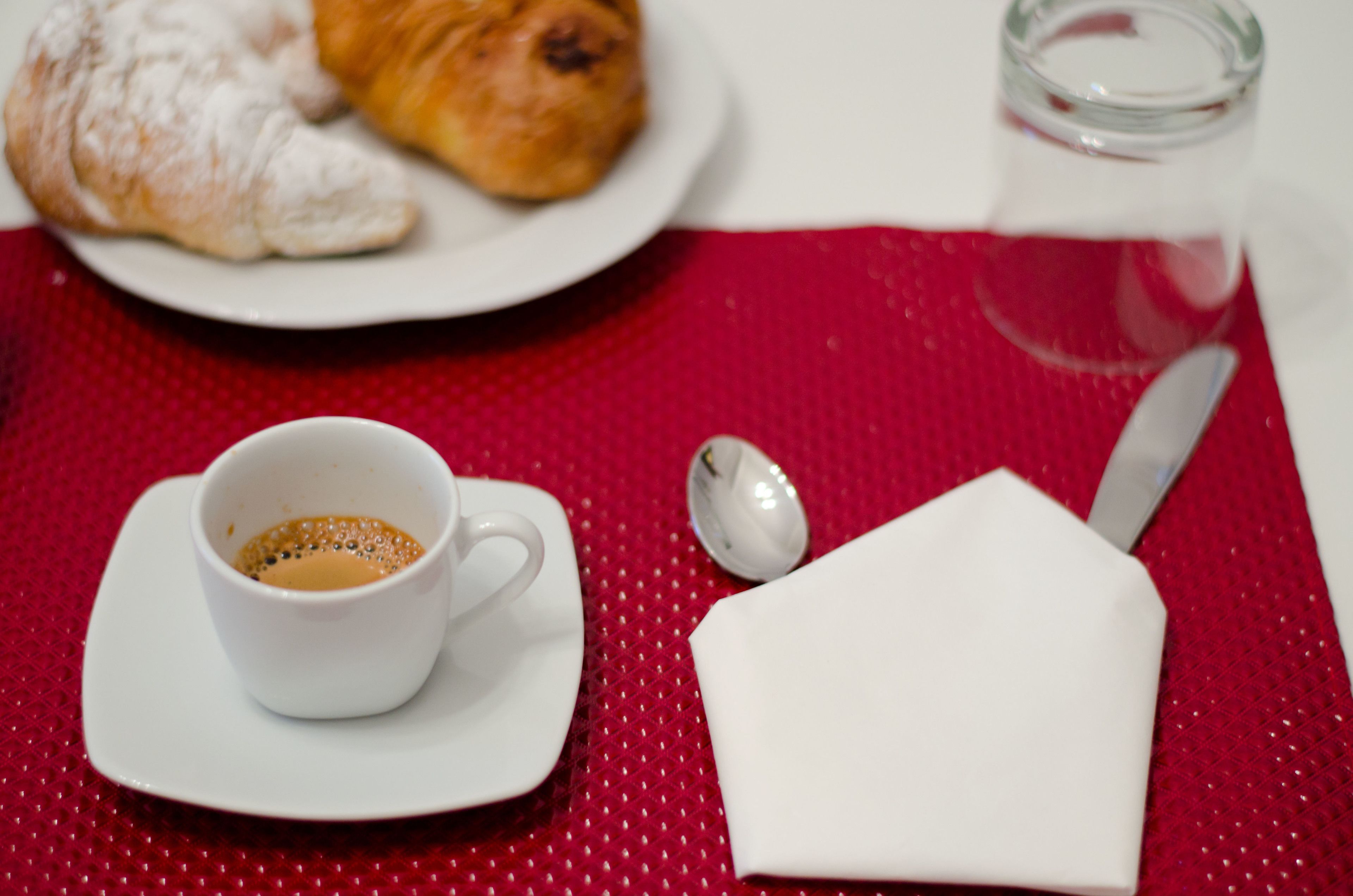 Una tavola apparecchiata con una tazza di caffè, un pasticcino, un cucchiaino e un tovagliolo su un sottopiattino rosso.