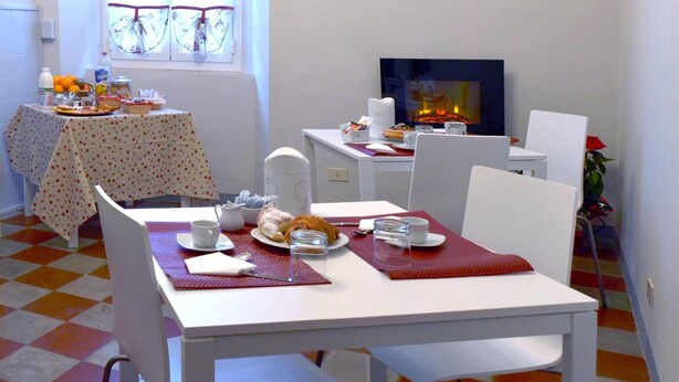 Un comedor con piso a cuadros, mesas y sillas blancas. Se ve una ventana con cortinas, una mesa con comida y bebidas, y una chimenea encendida.