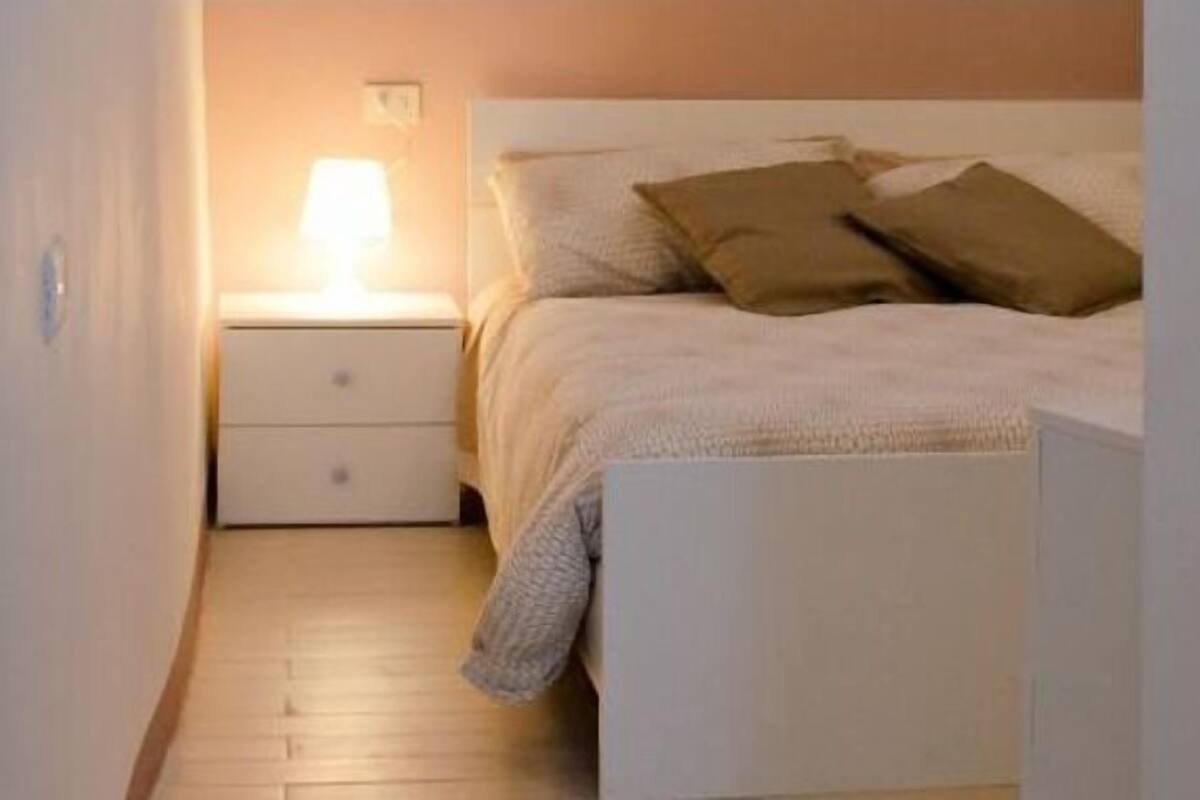 A bedroom with a bed, a nightstand with a lamp, and a wooden floor.