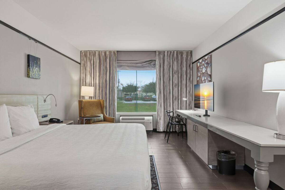 A hotel room with a large bed, a desk with a chair, a lamp, and a view of the outdoors.