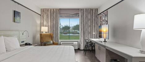 A hotel room with a large bed, a desk with a chair, a lamp, and a view of the outdoors.