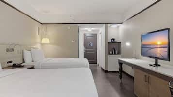 Habitación de hotel con dos camas, un escritorio con ordenador, un televisor mostrando un atardecer y una kitchenette.