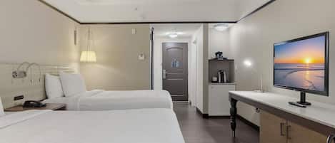 A hotel room with two beds, a desk with a computer, a TV displaying a sunset, and a kitchenette area.