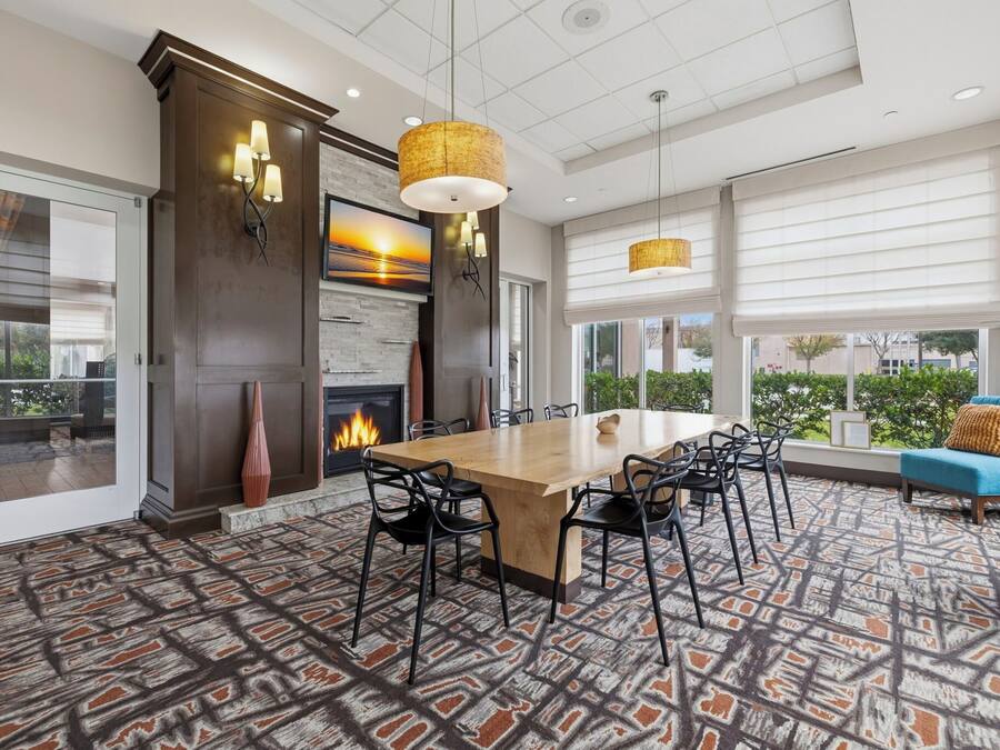 A spacious dining area with a large wooden table, black chairs, and a patterned carpet. There are two hanging pendant lights, a fireplace, and a large window with blinds.