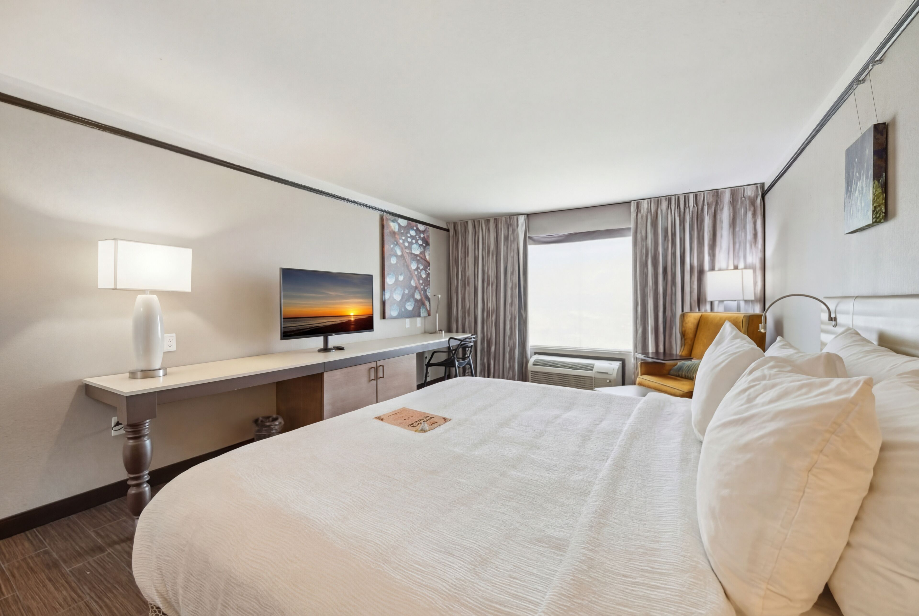 A hotel room with a large bed, a desk with a computer, a chair, a window with curtains, and a painting on the wall.