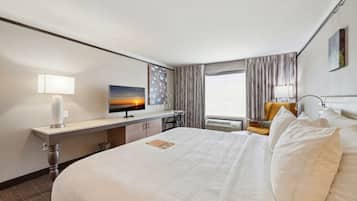 A hotel room with a large bed, a desk with a computer, a chair, a window with curtains, and a painting on the wall.