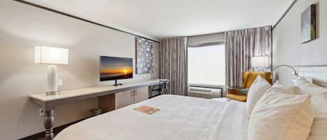 A hotel room with a large bed, a desk with a computer, a chair, a window with curtains, and a painting on the wall.