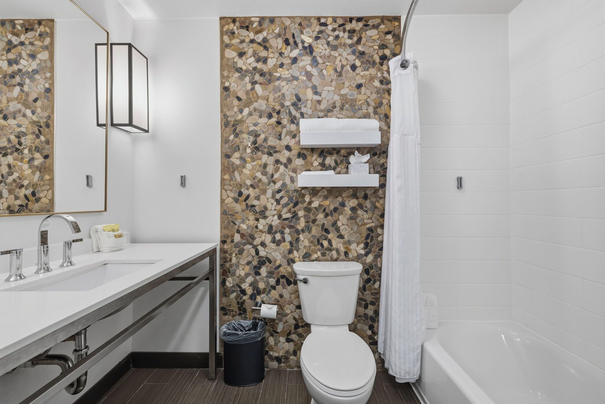 A modern bathroom with a pebble mosaic wall, a white toilet, a bathtub, and a vanity with a mirror and sink.