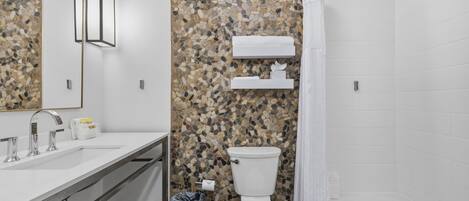 Een moderne badkamer met een mozaïekwand van kiezelstenen, een wit toilet, een badkuip en een wastafelmeubel met spiegel en wastafel.