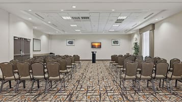 Une salle de conférence avec des rangées de chaises disposées face à un pupitre, un tapis à motifs et des tableaux encadrés accrochés aux murs.