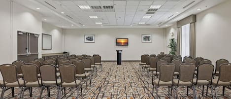 A conference room with rows of chairs facing a podium, a patterned carpet, and framed pictures on the walls.