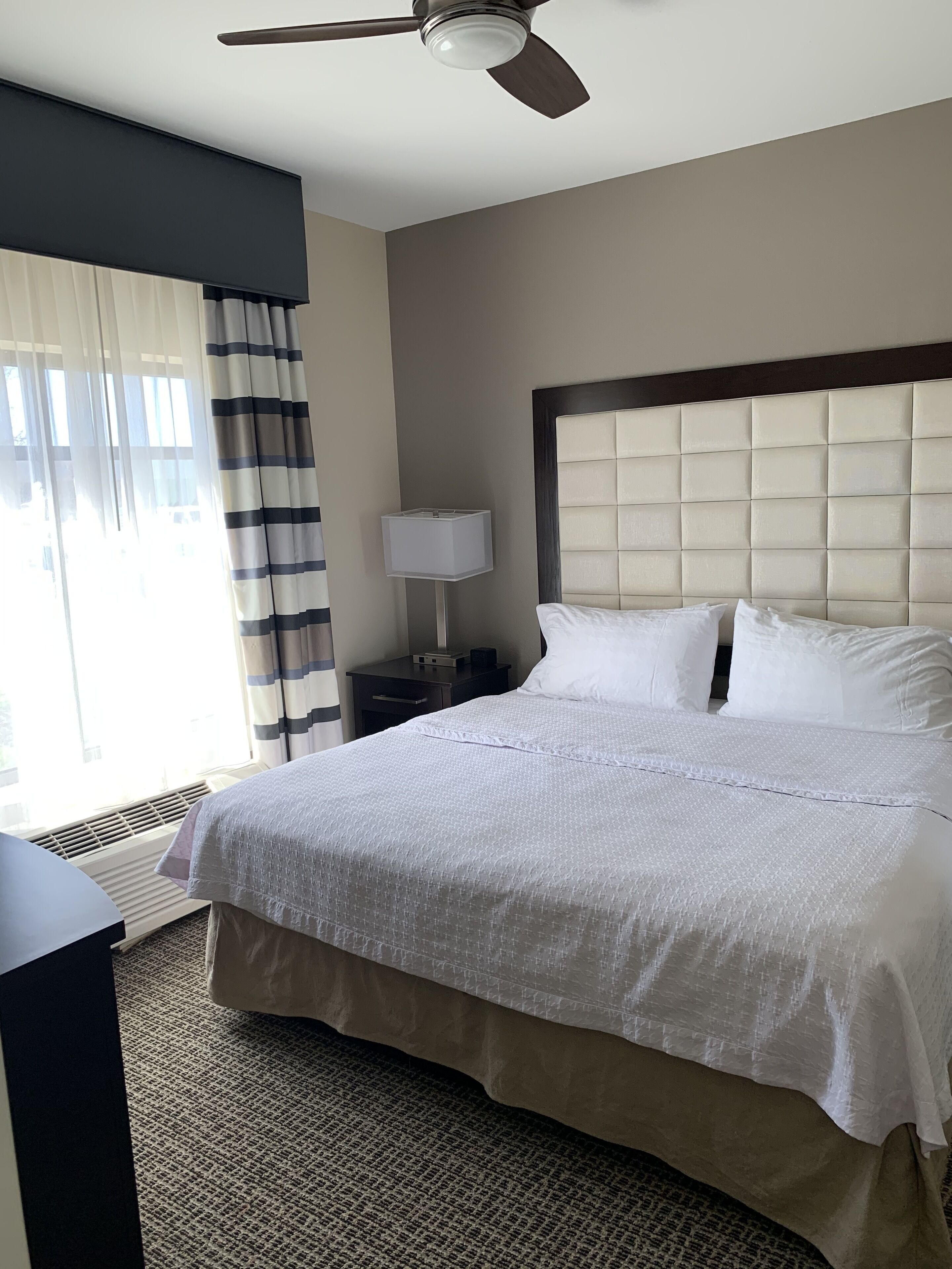 A hotel room with a large bed, a ceiling fan, a nightstand with a lamp, and a window with curtains.