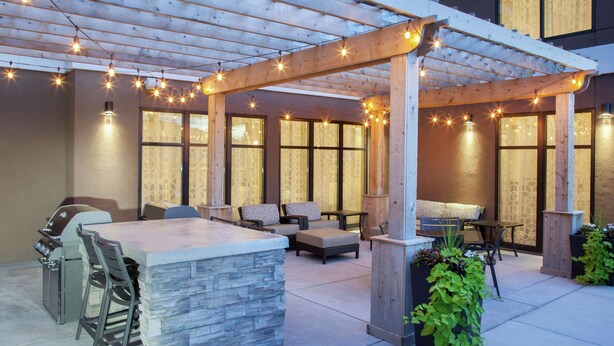An outdoor patio with a stone bar, wooden pergola, string lights, and seating. - Homewood Suites by Hilton Munster (Munster)