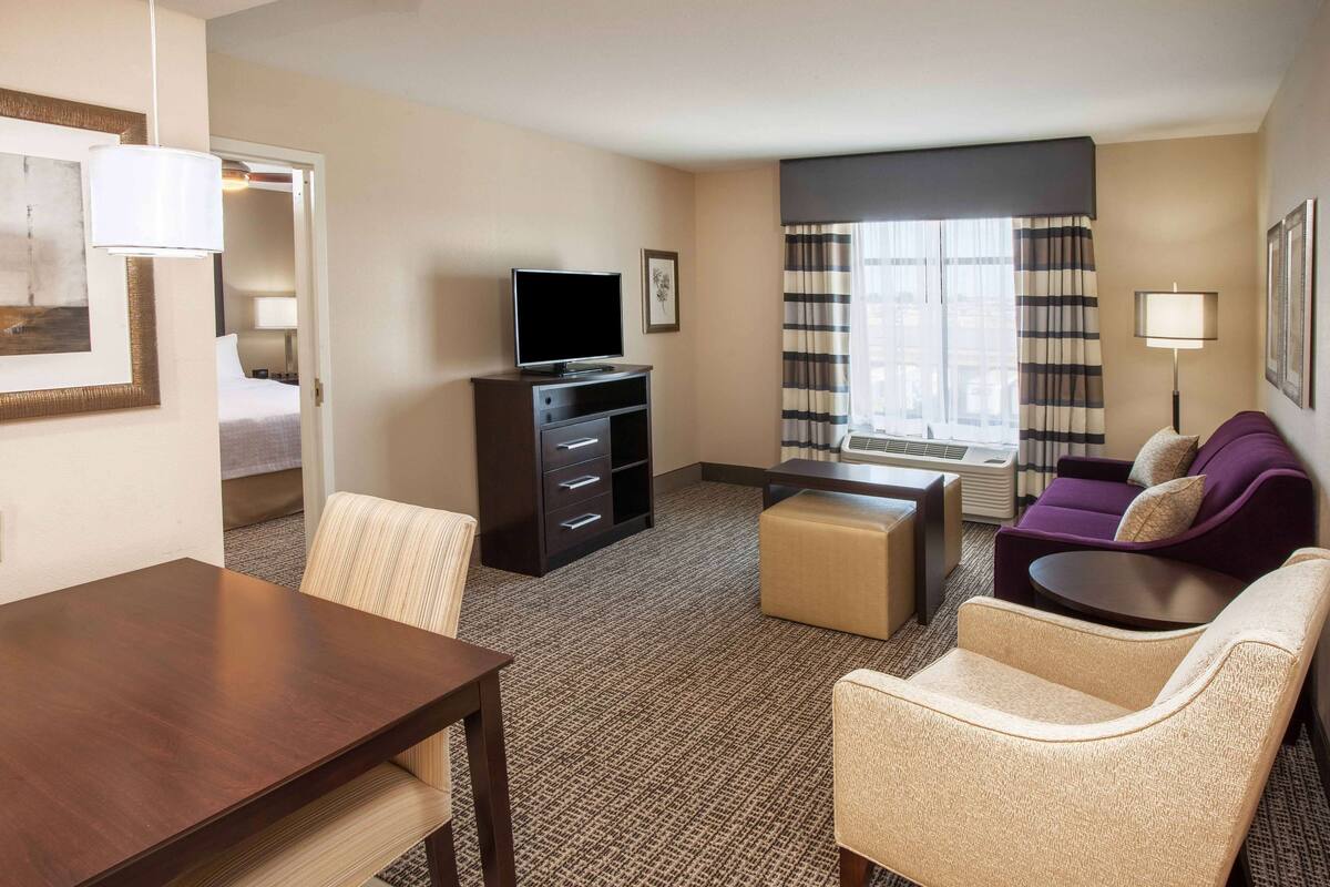 A hotel room with a bed, a TV on a dresser, a dining table, a sofa, and a window with curtains.