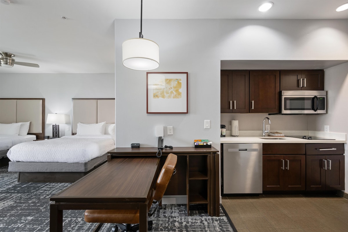 A hotel room with a bed, a desk, a chair, a dining table, a microwave, a sink, and cabinets.