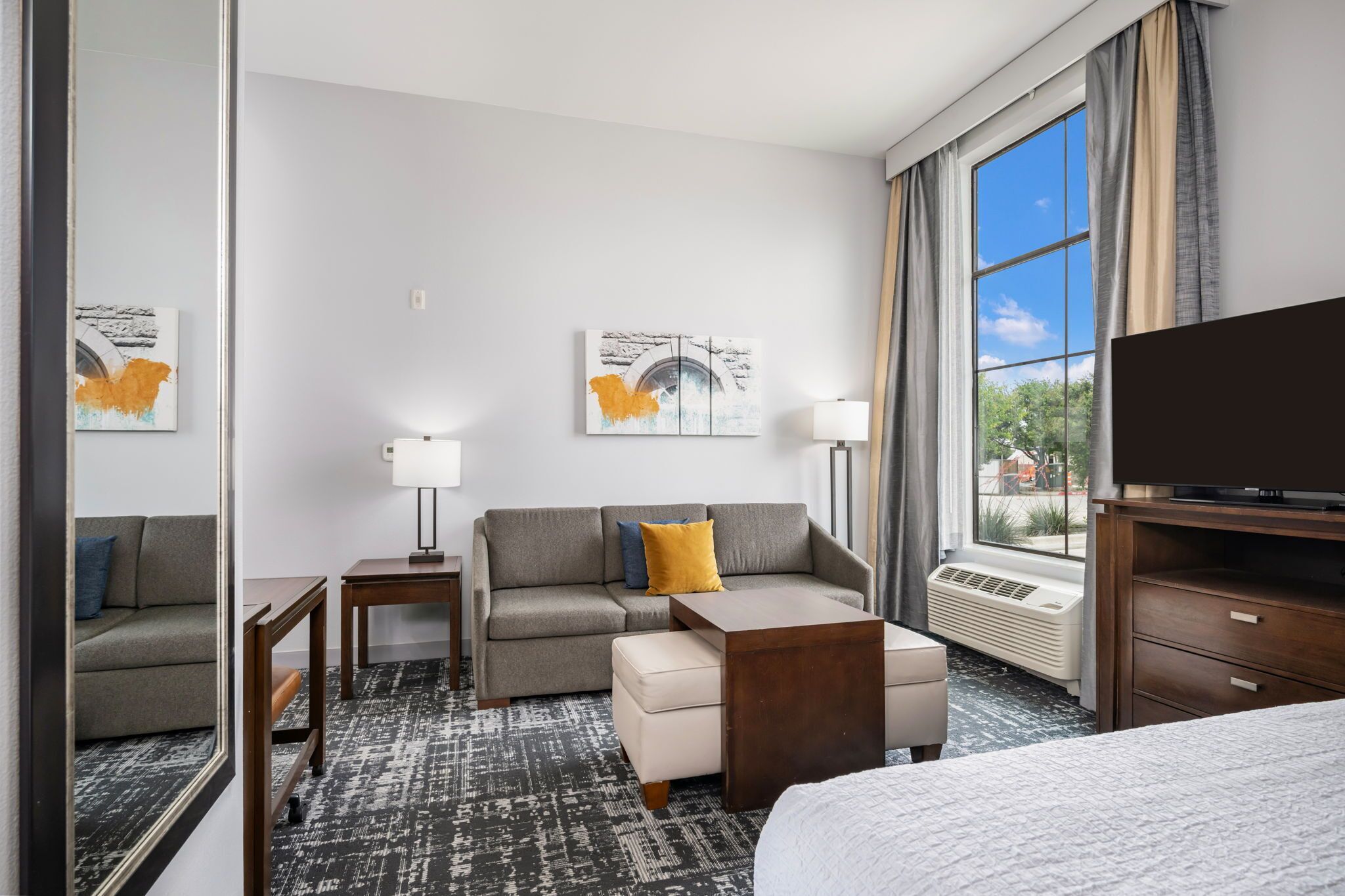 A hotel room with a sofa, ottoman, side table, lamp, TV, and a large window with curtains.
