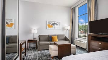 A hotel room with a sofa, ottoman, side table, lamp, TV, and a large window with curtains.
