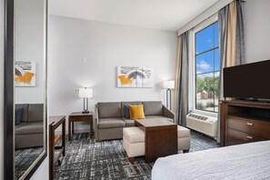 A hotel room with a sofa, ottoman, side table, lamp, TV, and a large window with curtains. - Homewood Suites By Hilton New Braunfels (New Braunfels)
