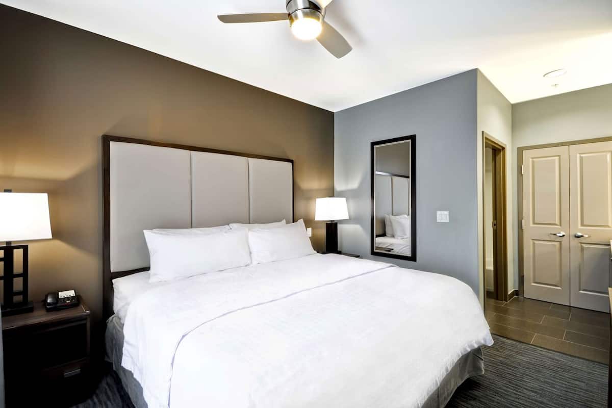 A hotel room with a bed, bedside lamps, a ceiling fan, a mirror, and a door.