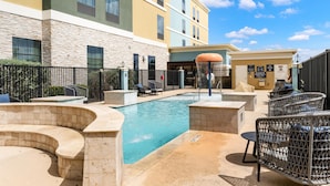 A pool area with lounge chairs, a covered seating area, and a building with multiple windows and a stone facade. - Homewood Suites By Hilton New Braunfels (New Braunfels)