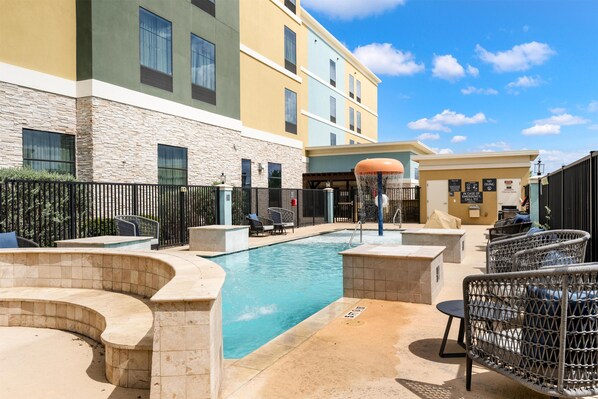 A pool area with lounge chairs, a covered seating area, and a building with multiple windows and a stone facade. - Homewood Suites By Hilton New Braunfels (New Braunfels)