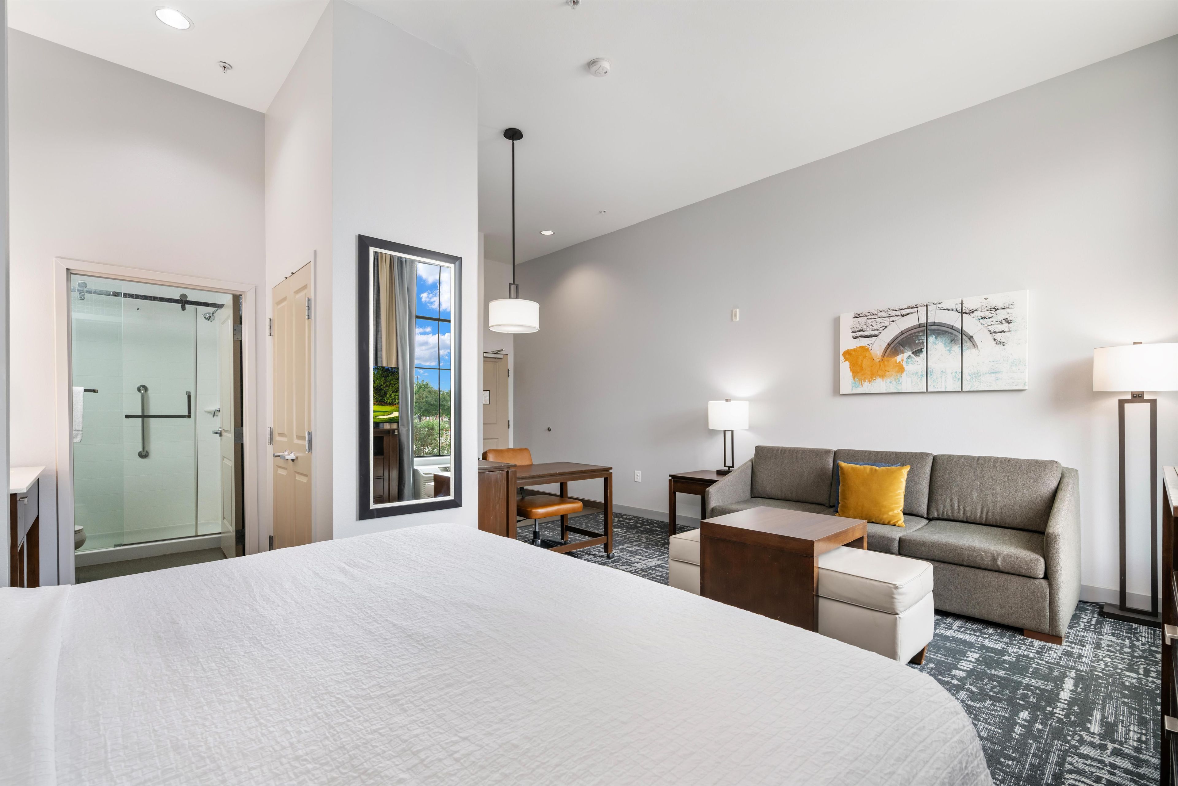 Habitación de hotel con una cama grande, un sofá, un escritorio, una silla, cabina de ducha de vidrio y un cuadro en la pared.