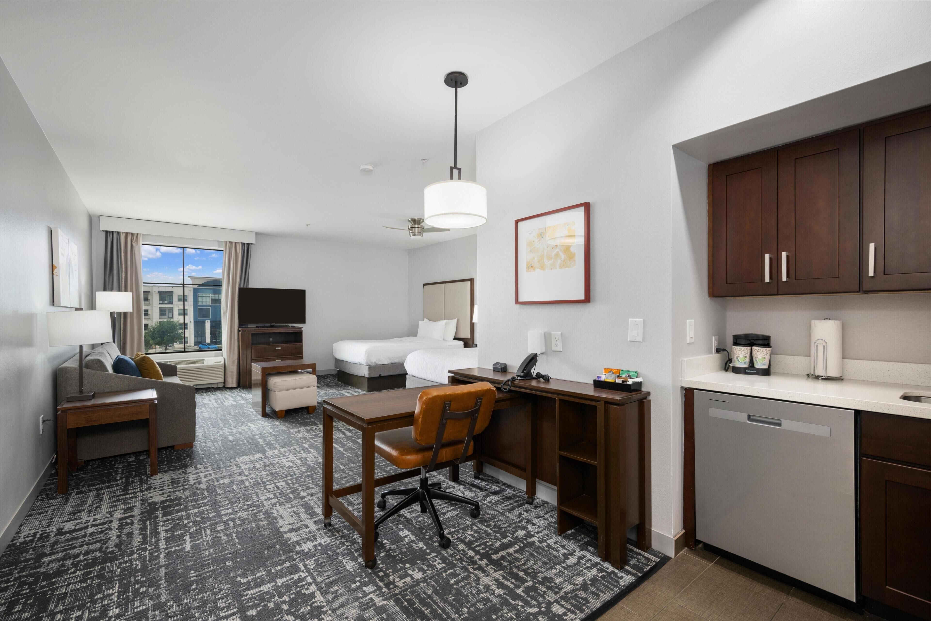 Ein Hotelzimmer mit Schreibtisch, Stuhl, Sofa und Bett. Es gibt ein Fenster mit Blick auf Gebäude, einen Deckenventilator und ein an der Wand befestigtes Kunstwerk.