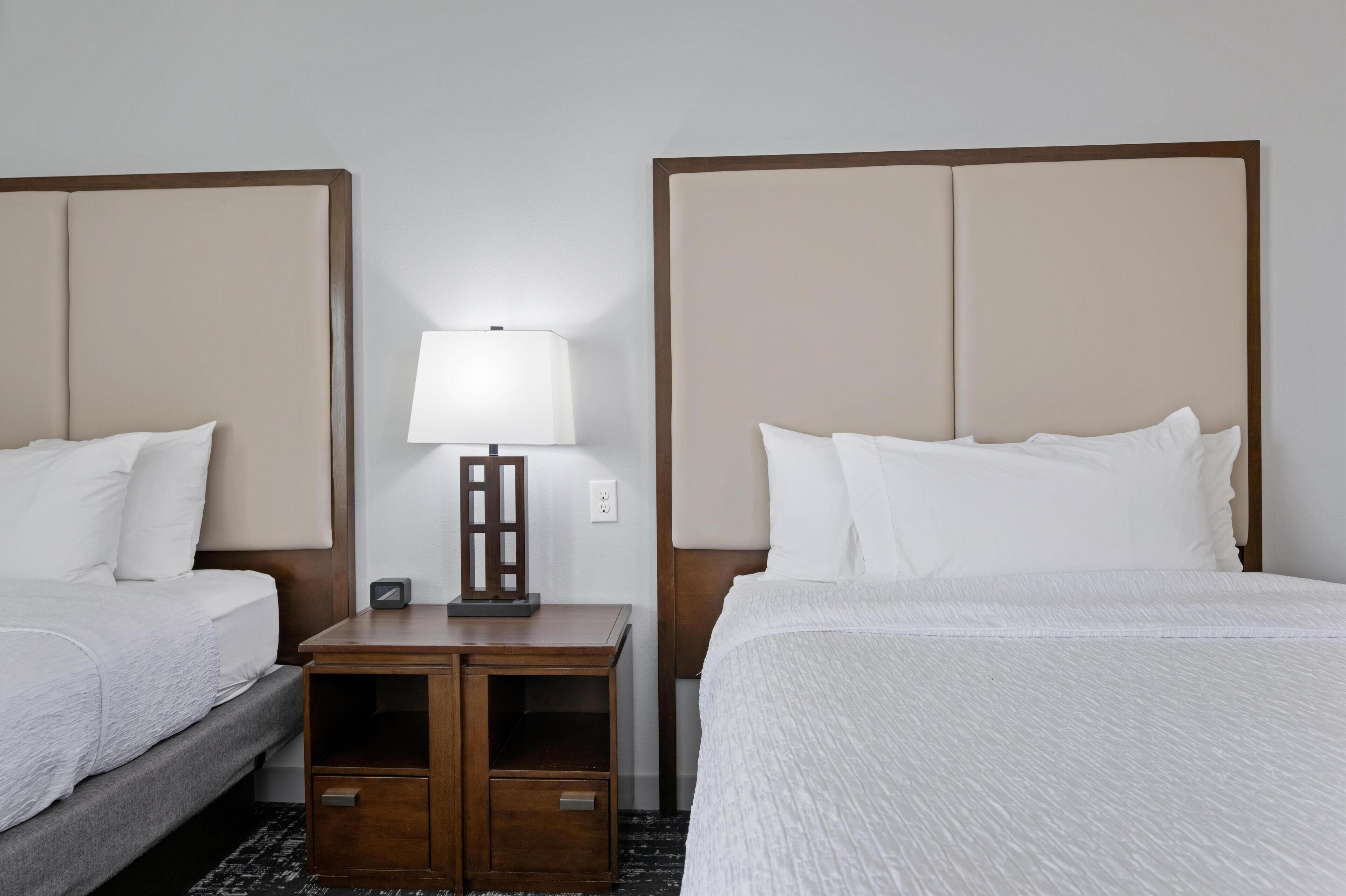 A hotel room with two beds, a nightstand with a lamp, and a small table.